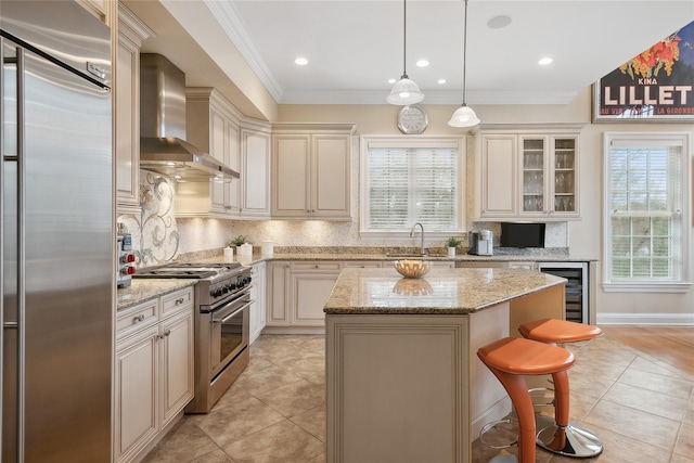 kitchen featuring wall chimney range hood, premium appliances, light stone counters, decorative light fixtures, and a kitchen island