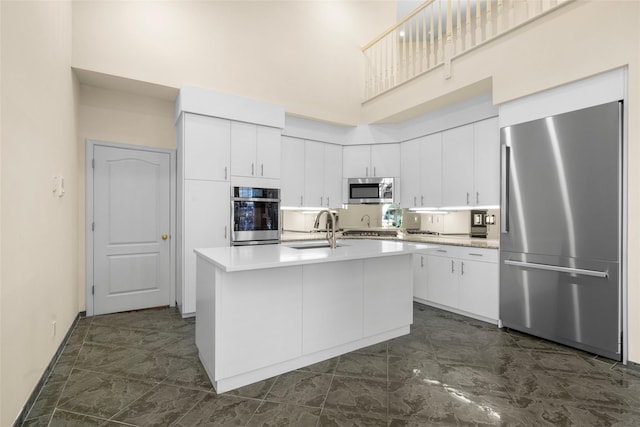 kitchen with sink, a towering ceiling, a kitchen island with sink, white cabinets, and appliances with stainless steel finishes