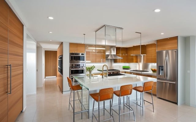 kitchen featuring stainless steel appliances, a large island with sink, wall chimney exhaust hood, and hanging light fixtures