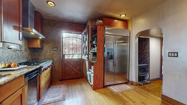 kitchen featuring wall chimney range hood, light stone countertops, tasteful backsplash, light hardwood / wood-style floors, and stainless steel appliances