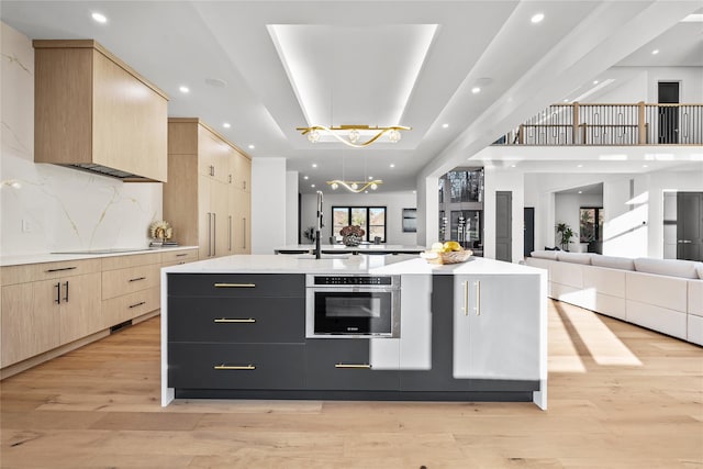 kitchen featuring stainless steel oven, hanging light fixtures, light hardwood / wood-style flooring, an island with sink, and light brown cabinetry