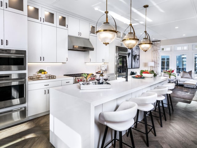 kitchen with white cabinets, dark parquet flooring, hanging light fixtures, and an island with sink