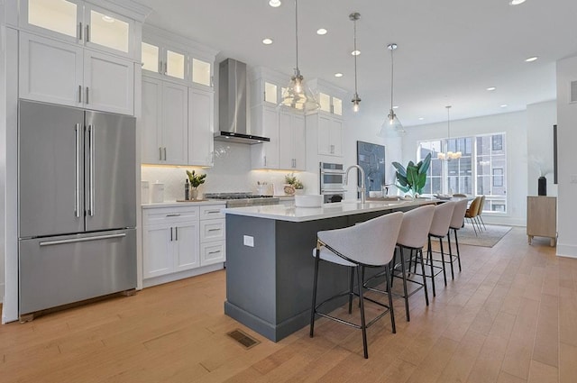 kitchen featuring a center island with sink, stainless steel appliances, light countertops, white cabinetry, and wall chimney range hood