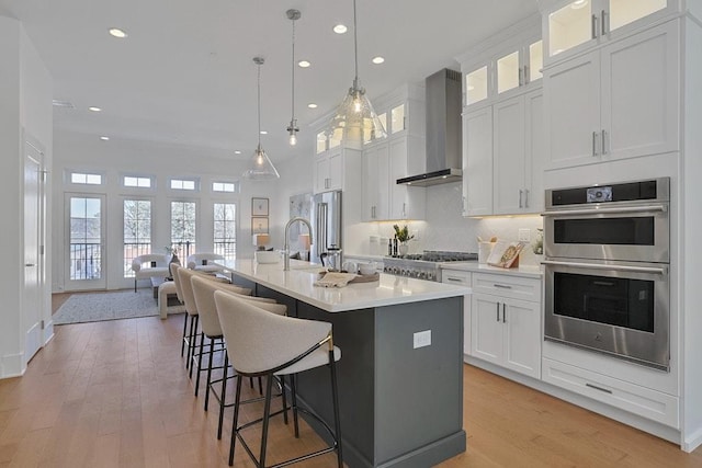 kitchen featuring wall chimney exhaust hood, a center island with sink, glass insert cabinets, and light countertops