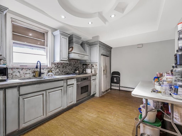 kitchen featuring appliances with stainless steel finishes, gray cabinetry, custom range hood, a tray ceiling, and light hardwood / wood-style floors