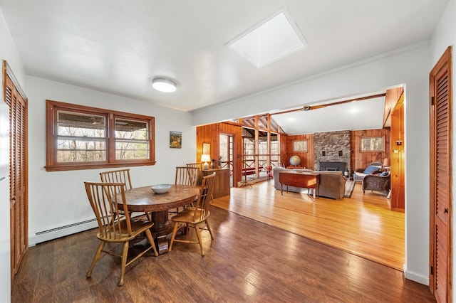 dining area featuring a skylight, wooden walls, a baseboard radiator, hardwood / wood-style floors, and a stone fireplace