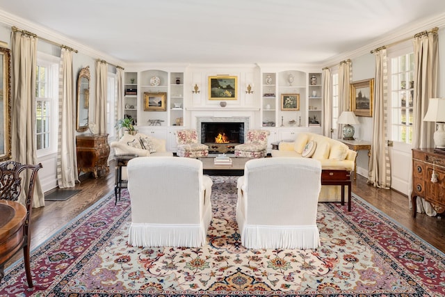 living room with a lit fireplace, built in shelves, ornamental molding, and dark wood-style flooring