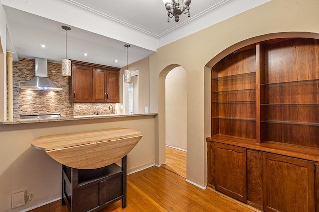 interior space with tasteful backsplash, light stone counters, wall chimney range hood, pendant lighting, and hardwood / wood-style floors