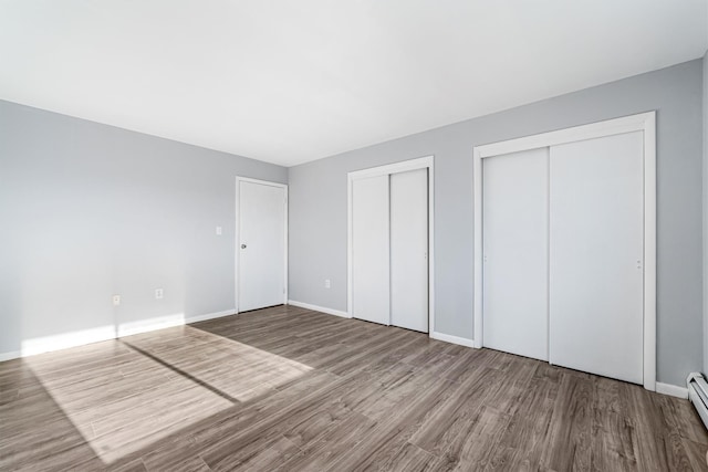 unfurnished bedroom featuring wood-type flooring, baseboard heating, and multiple closets