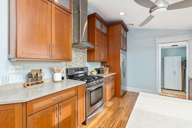 kitchen featuring light stone countertops, ceiling fan, stainless steel appliances, wall chimney range hood, and light hardwood / wood-style flooring