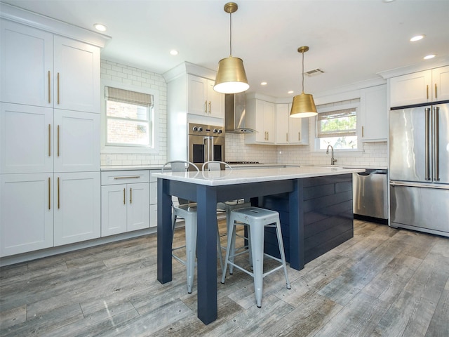 kitchen featuring appliances with stainless steel finishes, wall chimney range hood, light hardwood / wood-style flooring, white cabinetry, and hanging light fixtures