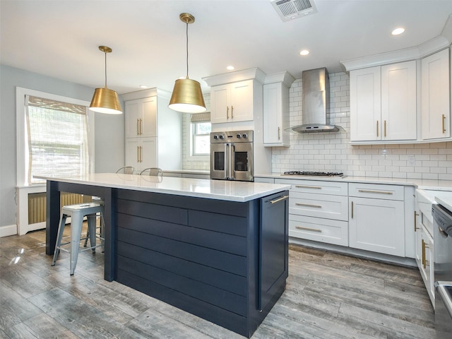 kitchen with pendant lighting, wood-type flooring, wall chimney range hood, white cabinetry, and stainless steel appliances