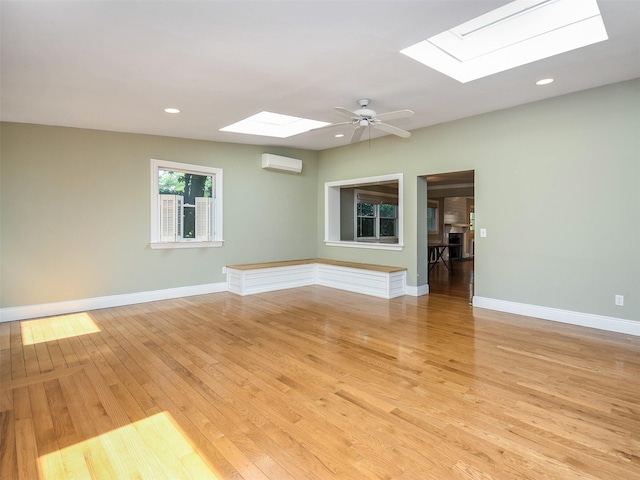 unfurnished room featuring a wall unit AC, a skylight, ceiling fan, and light hardwood / wood-style flooring