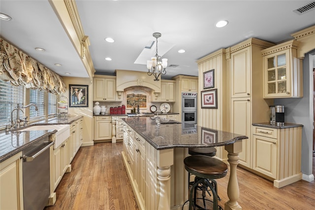 kitchen featuring appliances with stainless steel finishes, dark stone counters, a kitchen island with sink, pendant lighting, and cream cabinets