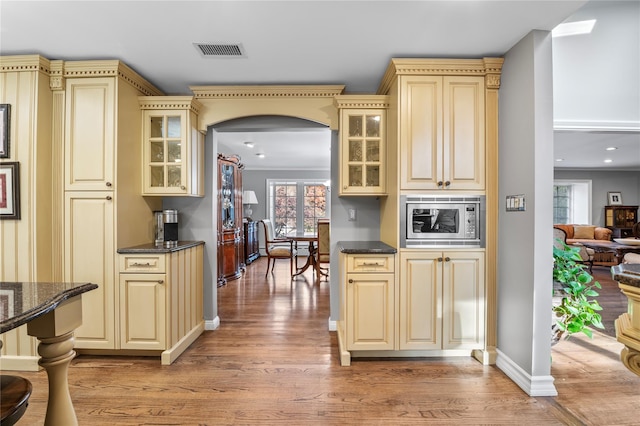 bar featuring stainless steel microwave, light hardwood / wood-style flooring, crown molding, dark stone counters, and cream cabinetry