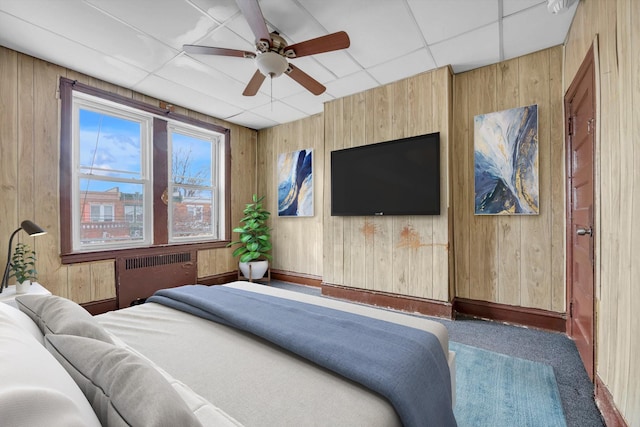 carpeted bedroom featuring a paneled ceiling, ceiling fan, radiator heating unit, and wooden walls