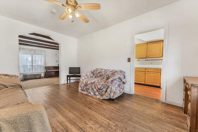 living area featuring radiator heating unit, a ceiling fan, wood finished floors, beamed ceiling, and baseboards