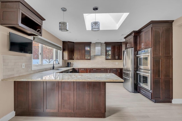 kitchen featuring a skylight, wall chimney exhaust hood, kitchen peninsula, pendant lighting, and appliances with stainless steel finishes