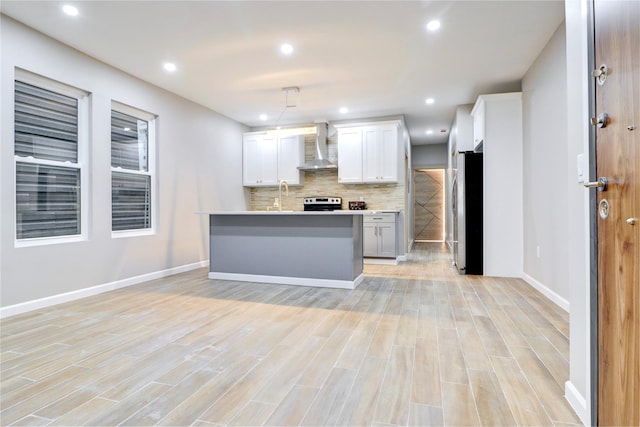 kitchen featuring appliances with stainless steel finishes, wall chimney exhaust hood, a kitchen island with sink, decorative light fixtures, and white cabinetry