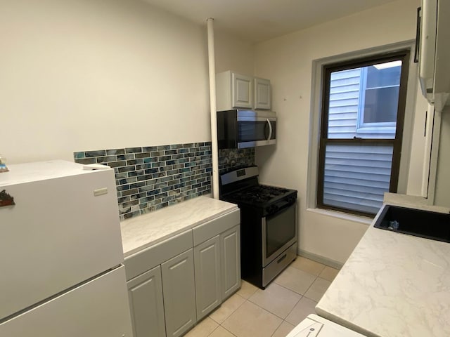 kitchen with gray cabinetry, backsplash, light tile patterned floors, white fridge, and gas stove