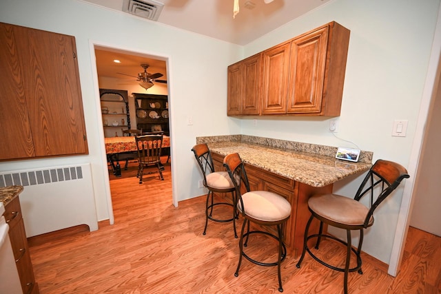 kitchen with light stone countertops, radiator, ceiling fan, light hardwood / wood-style floors, and a breakfast bar area