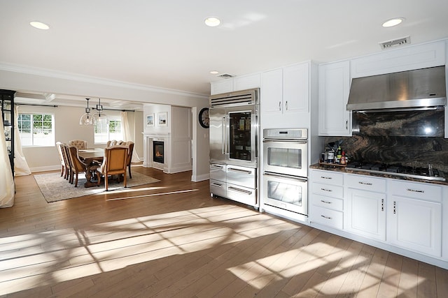 kitchen featuring ornamental molding, stainless steel appliances, wall chimney range hood, hardwood / wood-style flooring, and white cabinetry