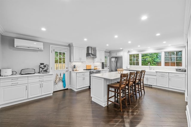 kitchen with appliances with stainless steel finishes, a wall mounted AC, wall chimney range hood, white cabinets, and a kitchen island