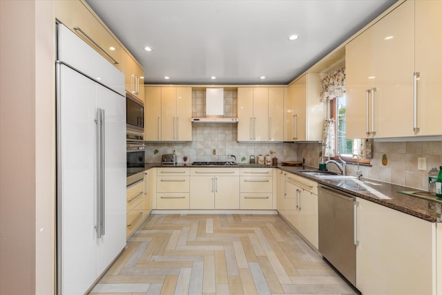 kitchen with sink, wall chimney exhaust hood, built in appliances, light parquet floors, and dark stone counters