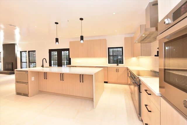 kitchen with light brown cabinets, a center island with sink, wall chimney range hood, decorative light fixtures, and a breakfast bar area