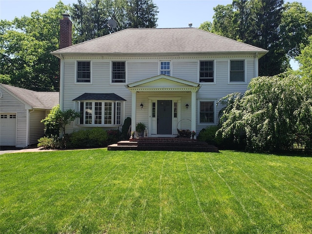 colonial house with an attached garage, roof with shingles, a chimney, and a front yard