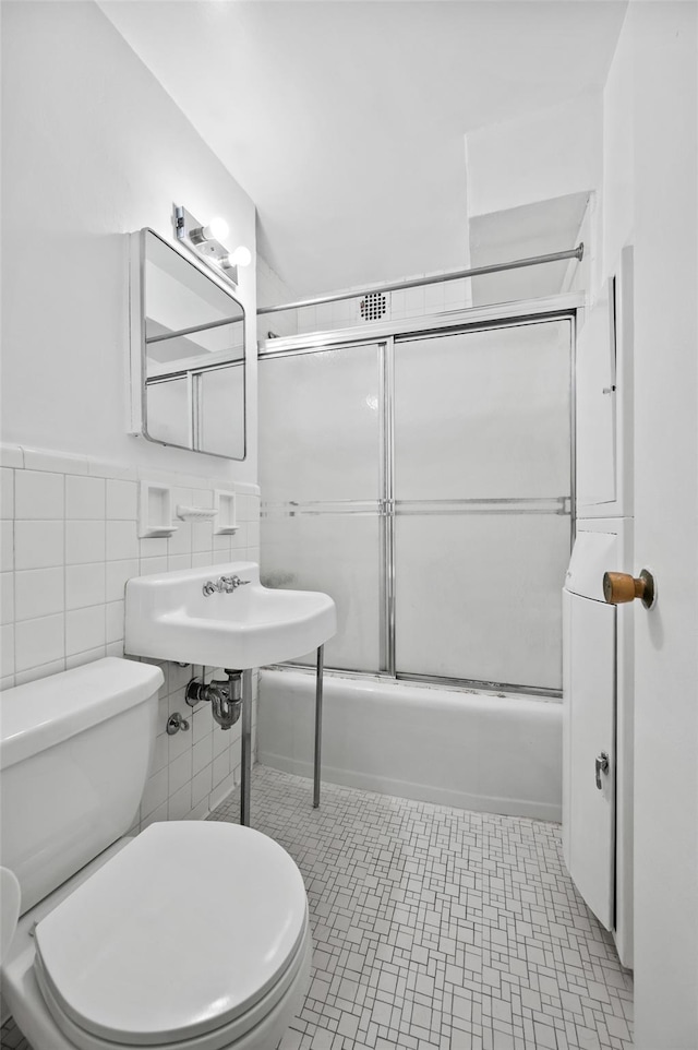 bathroom featuring toilet, shower / bath combination with glass door, and tile walls