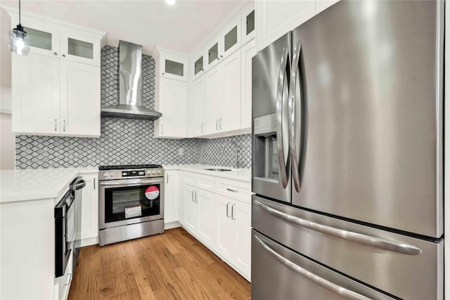 kitchen with white cabinets, hanging light fixtures, light hardwood / wood-style flooring, wall chimney exhaust hood, and appliances with stainless steel finishes