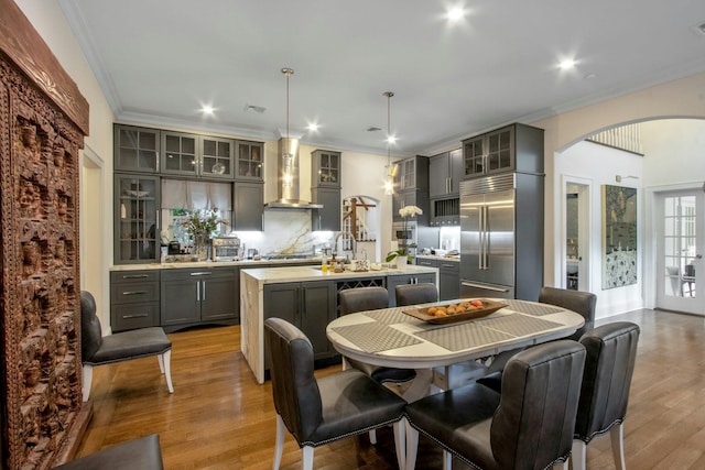 kitchen with gray cabinetry, wall chimney range hood, decorative light fixtures, a center island with sink, and appliances with stainless steel finishes