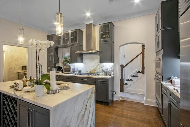 kitchen with pendant lighting, wall chimney exhaust hood, gray cabinets, ornamental molding, and an island with sink