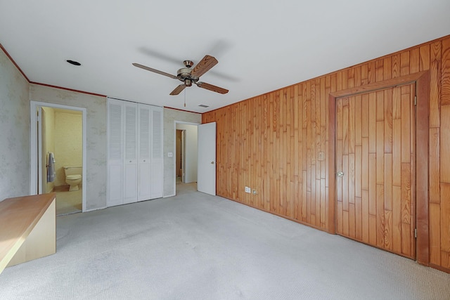 unfurnished bedroom featuring light colored carpet, ensuite bath, ceiling fan, and wood walls