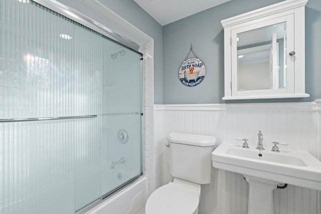 full bathroom with toilet, bath / shower combo with glass door, a sink, and wainscoting