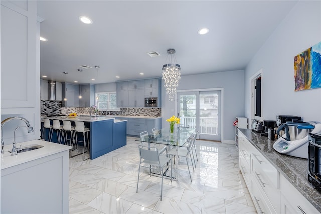 kitchen featuring a center island with sink, a kitchen breakfast bar, wall chimney range hood, sink, and hanging light fixtures