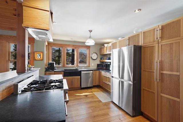 kitchen featuring stainless steel appliances, a sink, hanging light fixtures, light wood finished floors, and dark countertops