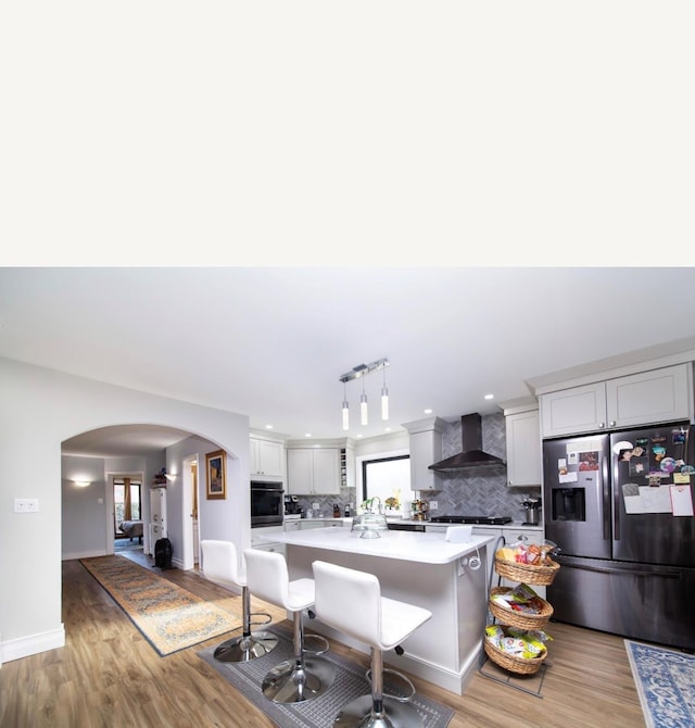 kitchen with wall chimney exhaust hood, stainless steel appliances, a kitchen breakfast bar, decorative light fixtures, and light wood-type flooring