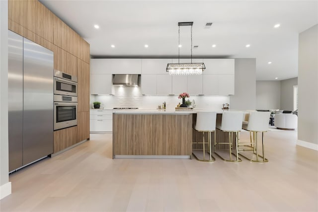 kitchen featuring white cabinetry, a center island with sink, appliances with stainless steel finishes, decorative light fixtures, and wall chimney exhaust hood