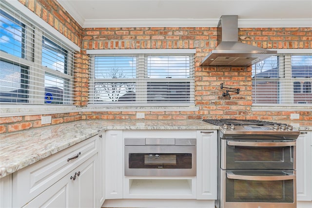 kitchen with wall chimney exhaust hood, light stone countertops, stainless steel range, white cabinetry, and brick wall