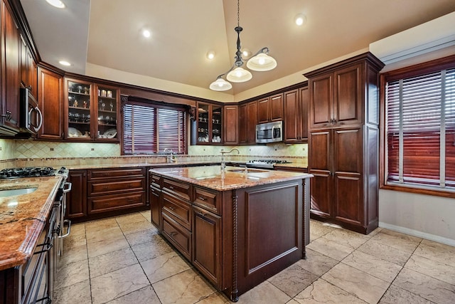 kitchen featuring light stone countertops, stove, tasteful backsplash, pendant lighting, and a center island with sink