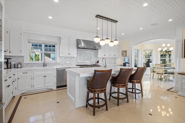 kitchen featuring stainless steel dishwasher, wall chimney range hood, pendant lighting, white cabinets, and an island with sink