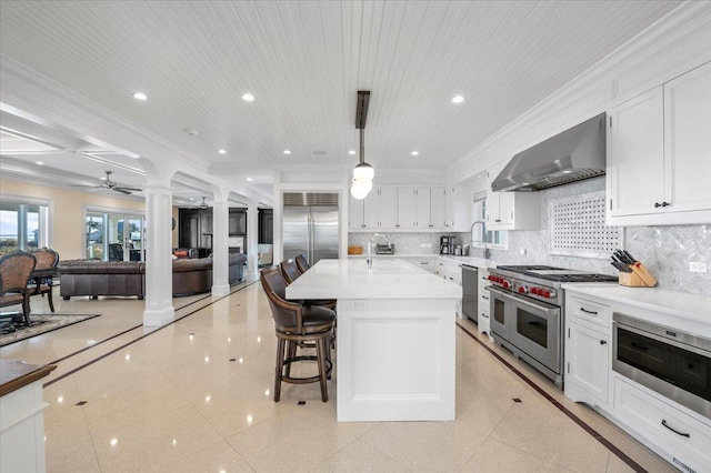 kitchen featuring ceiling fan, wall chimney range hood, pendant lighting, high quality appliances, and an island with sink