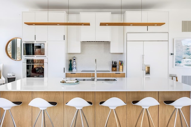 kitchen featuring white cabinetry, tasteful backsplash, built in appliances, decorative light fixtures, and a kitchen island with sink