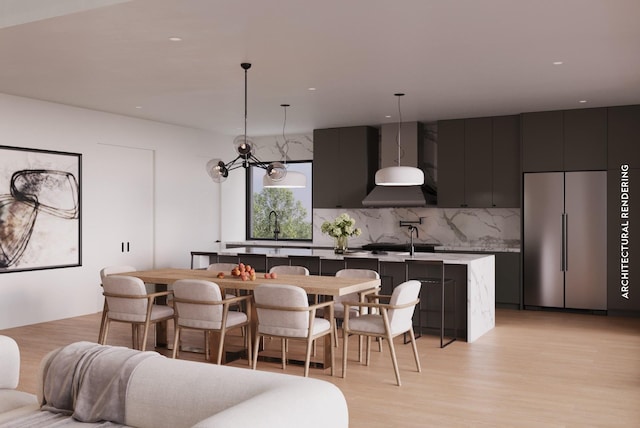 kitchen featuring wall chimney exhaust hood, decorative backsplash, an island with sink, decorative light fixtures, and stainless steel built in refrigerator