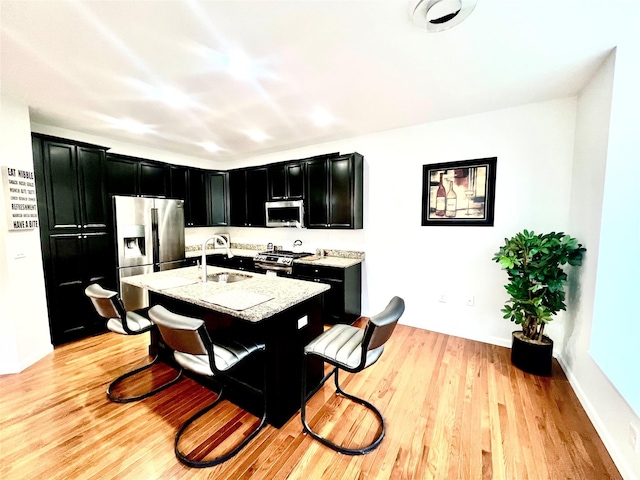 kitchen featuring a center island with sink, appliances with stainless steel finishes, light hardwood / wood-style floors, light stone countertops, and sink