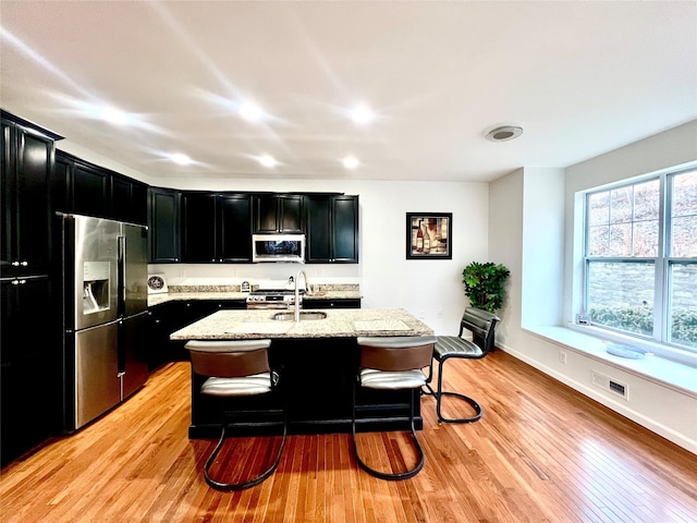 kitchen with a breakfast bar area, a center island with sink, light hardwood / wood-style floors, stainless steel appliances, and light stone countertops