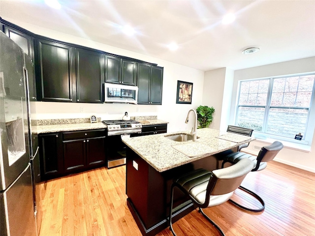 kitchen with an island with sink, appliances with stainless steel finishes, light stone countertops, and light hardwood / wood-style flooring