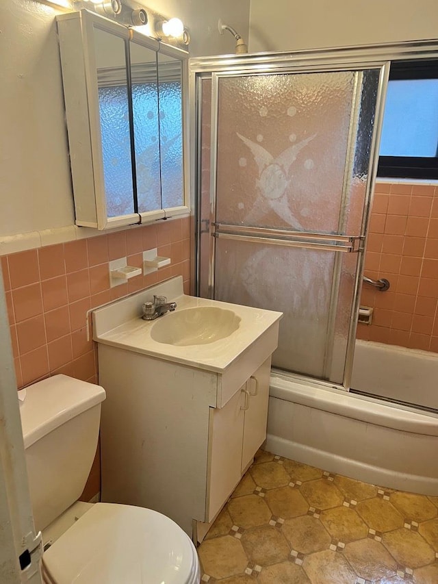 full bathroom with combined bath / shower with glass door, vanity, toilet, and tile walls